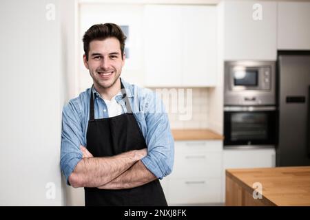 handsome young man smiling in the kitchen wearing an apron - Portrait of a cheerful confident caucasian young man looking at camera with arms crossed  Stock Photo