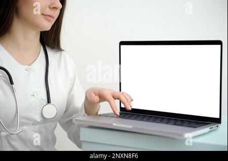 Female medical assistant wears white coat, headset video calling distant patient on laptop. Doctor talking to client using virtual chat computer app. Telemedicine, remote healthcare services concept Stock Photo