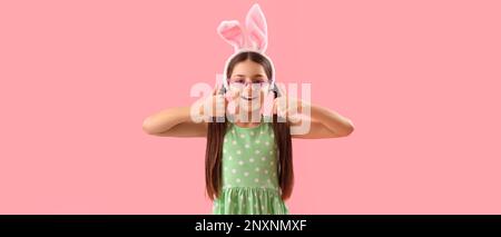 Cute little girl with bunny ears showing thumb-up on pink background Stock Photo