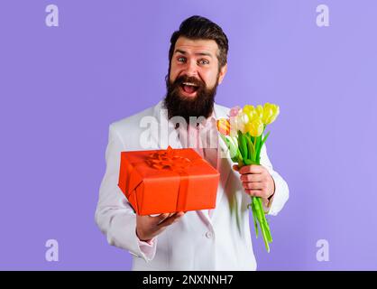 Smiling bearded man with gift box and bouquet of flowers. Businessman in suit with gift and bouquet of tulips. Romantic man with bouquet of flowers Stock Photo