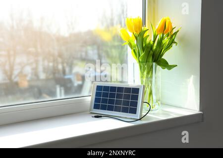 Portable solar panel and vase with tulips on windowsill in room Stock Photo