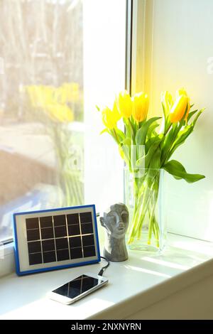 Portable solar panel charging mobile phone, decor and vase with tulips on windowsill in room Stock Photo