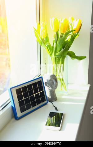 Portable solar panel charging mobile phone, decor and vase with tulips on windowsill in room Stock Photo