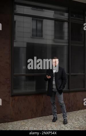 Serious young man in black coat is standing on streetand carefully studying information on his phone. Freelance.  Stock Photo