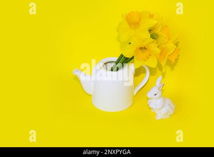 Jug with beautiful Daffodils and Easter bunny on yellow background with space for text. Home interior with easter decor. Daffodils in a vase, spring f Stock Photo