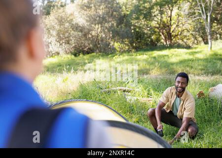 Happy, diverse couple camping in forest, over shoulder view of man pitching tent, copy space Stock Photo