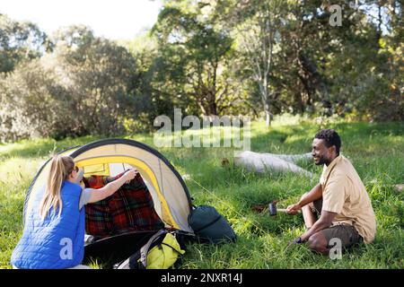 Happy, diverse couple camping in forest, pitching tent, setting up camp Stock Photo