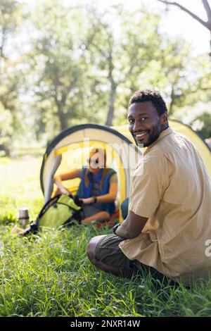 Vertical of happy, diverse couple camping in forest, setting up tent, man smiling, copy space Stock Photo