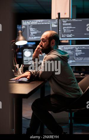 IT engineer answering landline phone call in office, talking about coding html script on cord telephone line. Working late at night to develop new computer system and app servers. Stock Photo