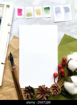 Watercolor paper sheet mockup with Christmas decor, brush, paints on a marble background. Mockup for presentation of illustrations, prints, logos, pos Stock Photo