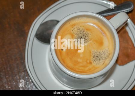 Freshly brewed coffee crema in a white cup with biscuit and spoon on a wooden table, view from above, copy space, selected focus, very narrow depth of Stock Photo