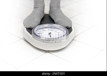 Feet in gray socks standing on a personal scale on the white tiled bathroom floor for weight measuring, copy space, selected focus Stock Photo