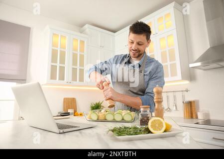 Man adding spices to dish while watching online cooking course via laptop in kitchen Stock Photo