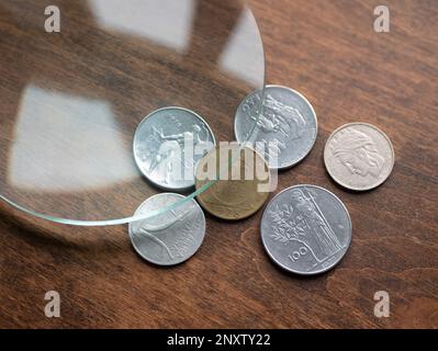 Old Italian Lira coins under magnifying glass on a wooden table, selective focus. Local currencies that ceased to exist Stock Photo