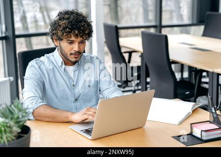 Focused smart confident guy of Indian nationality with curly hair, a programmer, IT specialist, sits at a table in the office, works in a laptop, works on a new program, at software support Stock Photo