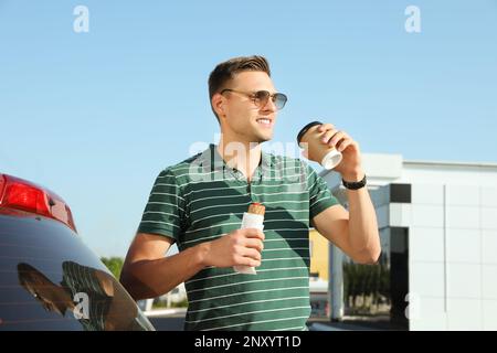 Young man with hot dog drinking coffee near car at gas station Stock Photo