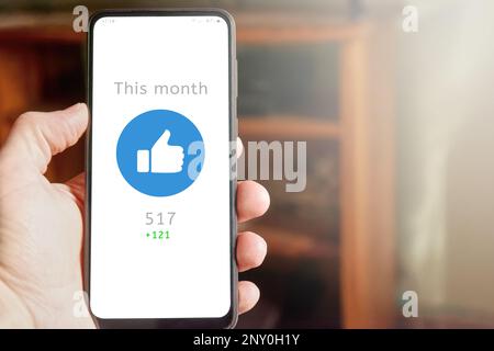 Hand holding smartphone with thumb up icon on the screen, social media report showing positive green statistics of engagement. Community interaction, Stock Photo