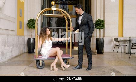 Elegant couple at entrance to hotel. Action. Elegant woman sits on European luggage trolley in hotels. Beautiful luxury couple at expensive hotel. Stock Photo