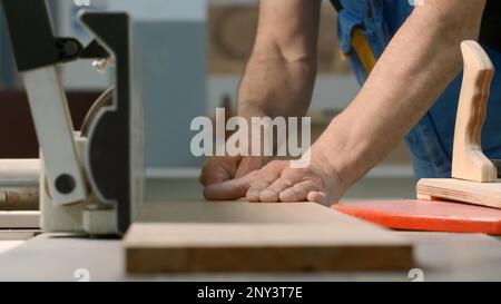 Close-up of man cutting board on machine. Creative. Carpenter processes wooden board on machine. Industrial processing and trimming of wooden boards w Stock Photo