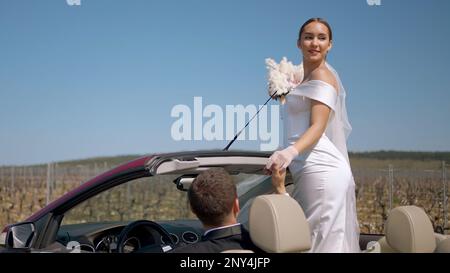 A young bride looks out of the car. Action. A model in a white dress with red lips holds a wedding bouquet in her hands and poses against a blue sky. Stock Photo
