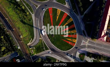 Aerial view of a round shaped crossroad with moving cars. Clip. Green meadow and bright growing flowers creating triangular pattern Stock Photo