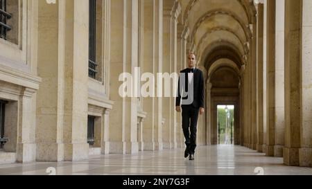Man in black and white official suit walking in arcuate shaped hall. Action. Man walking outdoors by the building with columns Stock Photo