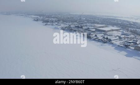 Aerial view of a town located by the lake in a morning haze. Clip. Village covered by snow, frozen river Stock Photo