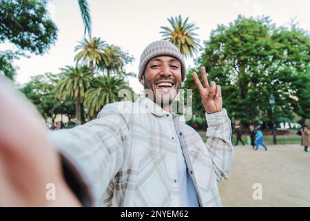 Young handsome man smiling taking a selfie portrait with a smartphone doing the peace sign with the fingers in a park outdoors wearing a beanie hat. Lifestyle concept. High quality photo Stock Photo