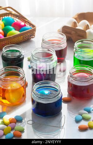 Dyeing Easter Eggs in Colorful Dye in Glass Jars with Multicolored Easter Candy on Marble Counter Stock Photo