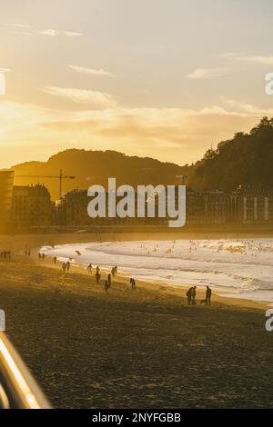 People strolling on sandy beach near waving sea against mountains and buildings in evening time during sunset Stock Photo