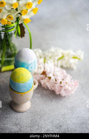Easter card concept with colorful eggs on eggcups near seasonal flowers on concrete background Stock Photo