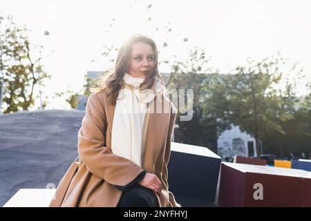 Contemplating female model in stylish coat and scarf smiling and looking away while relaxing on play ground and admiring cityscape Stock Photo