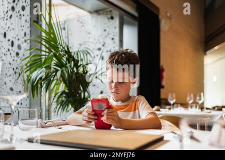 Concentrated boy in striped t shirt sitting at table with glasses and silverware while browsing smartphone in modern cafe Stock Photo