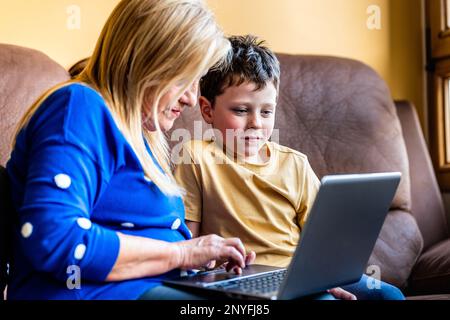 Positive mature grandmother smiling while sitting on comfortable sofa with adorable grandchild and surfing laptop together Stock Photo