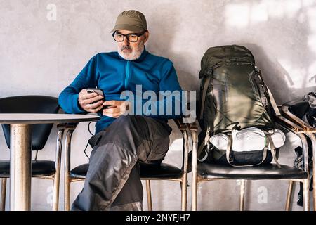 Concentrated bearded male traveler in casual clothes and eyeglasses with cap sitting at table near backpack while messaging on mobile phone during tri Stock Photo