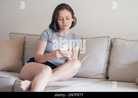 Concentrated young female in domestic clothes sitting on sofa while browsing smartphone at home Stock Photo
