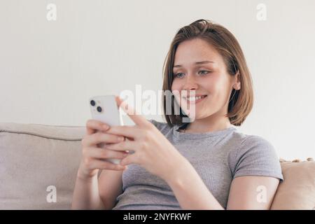 Concentrated young female in domestic clothes sitting on sofa while browsing smartphone at home Stock Photo