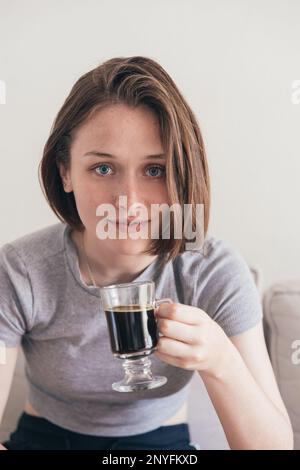 Positive young female in casual clothes sitting on sofa with cup of coffee while looking at camera Stock Photo