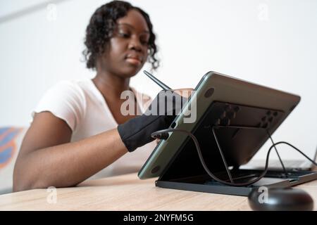 From below focused African American female in glove using stylus while drawing on graphic tablet at wooden table Stock Photo