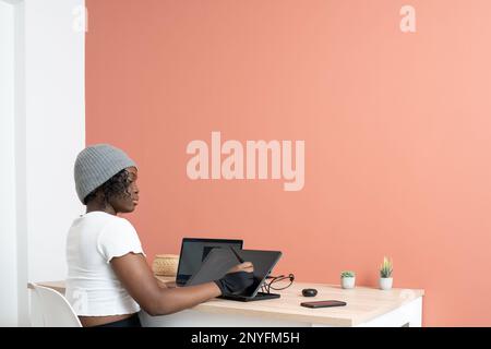 Back view of focused young African American female designer in white t shirt and hat sitting at table with stylus while drawing on graphic tablet Stock Photo