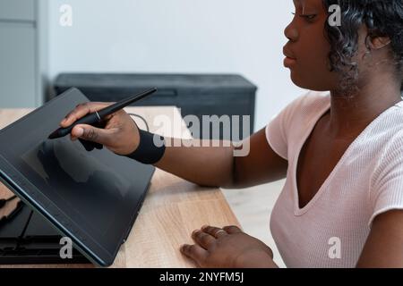 Side view of focused African American female in glove using stylus while drawing on graphic tablet at wooden table Stock Photo