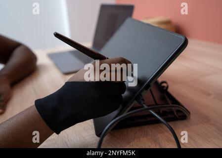 Unrecognizable African American female in glove using stylus while drawing on graphic tablet at wooden table Stock Photo
