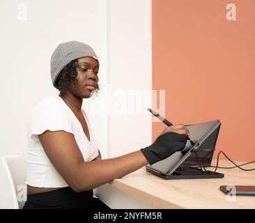 Side view of focused African American female in glove using stylus while drawing on graphic tablet at wooden table Stock Photo