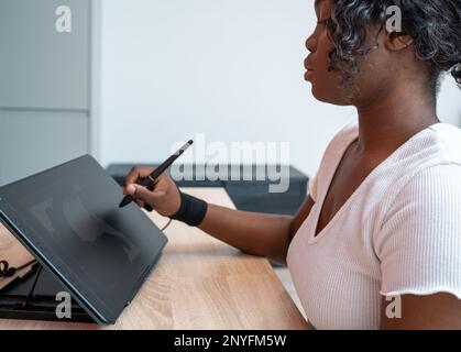 Side view of focused African American female in glove using stylus while drawing on graphic tablet at wooden table Stock Photo