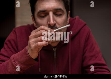 Crop relaxed male in red hoodie with closed eyes smelling aromatic blunt with cannabis leaves Stock Photo