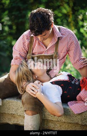 Lovely woman in traditional Austrian outfit lying on knees of handsome guy while relaxing in garden on sunny day Stock Photo