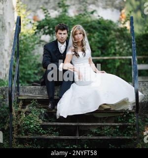 Young bride and groom in wedding costumes looking at camera while sitting and standing on steps near ancient building Stock Photo