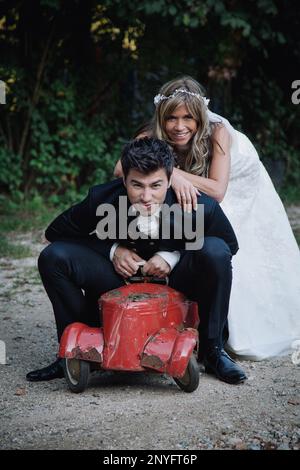 Attractive young bride and groom smiling and looking at camera while playing with ancient toy car in garden Stock Photo