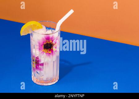 Transparent glass of refreshing cold drink decorated with pink flowers and straw placed on blue surface against bright orange wall Stock Photo