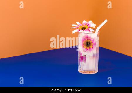 Transparent glass of refreshing cold drink decorated with pink flowers and straw placed on blue surface against bright orange wall Stock Photo